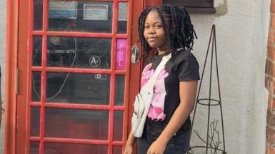 A girl is smiling while standing next to a red phone box. She has a black and pink Barbie T-shirt, jeans and a white bag.