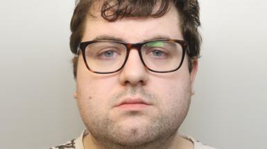 A custody image of Nathan Bennett. He has brown glasses on, a neutral expression, and mid-length brown hair. He is wearing a cream T-shirt with a brown, leaf-life design on it.