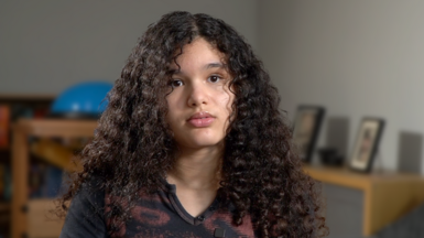 Mia looks into the camera with a neutral expression. She has curly dark coloured hair that just passes her shoulders. She is wearing a dark coloured top. In the background is living room furniture and photographs are propped on one side. 