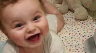 Baby Preston Davey smiles at the camera dressed in a white vest and appears to be lying in a cot with star bedsheet cover