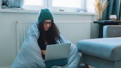 Women in a woolly hat and wrapped in a duvet sits next to a radiotor