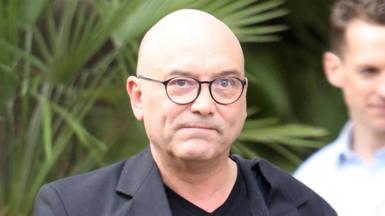 Gregg Wallace, wearing black round glasses with a black T-shirt and black jacket. He is looking pensive.