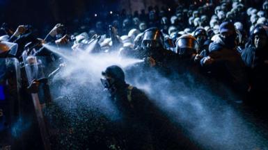 Police spray gas at protesters in Istanbul. It is dark with many people in shot. 