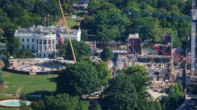 An aerial image of the White House, a large white building with tall classic columns. To the right of the building there's a large construction site, with scaffolding and cranes building a new structure. 