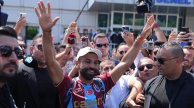 Mohamed Salah smiling with his hands in the air in a Trabzonspor shirt while being surrounded by people outside the airport