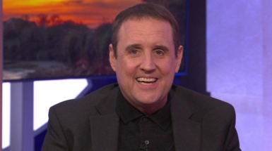 Peter Kay is wearing a black polo shirt under a black jacket and smiling at the camera. 
