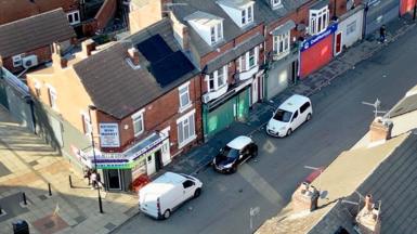 An aerial view of a residential street lined with brick terraced houses. Several vehicles, including white vans and small cars, are parked along the road. On the corner, a shop with signage reading “Hexthorpe Mini Market” stands at the junction. Rooftops with chimneys and solar panels are visible in the foreground, and a mostly empty street extends into the distance.