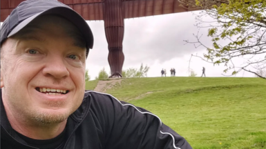 A man in a black baseball cap and black top smiling at the camera. He is outside and there is grass in the background.