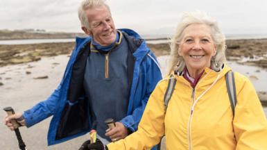 Stock photo of an older couple walking on the beach smiling at the camera looking natural and happy. A man is wearing a blue anorak and a woman is wearing a yellow anorak. They are both holding hiking sticks 