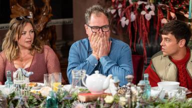 Alan Carr holds his hands to his mouth in surprise, while wearing a blue denim shirt. Two celebrities are pictured either side of him at the breakfast table in The Celebrity Traitors, with decorative table flowers, plates, bowls and a teacup in front of them on the table.
