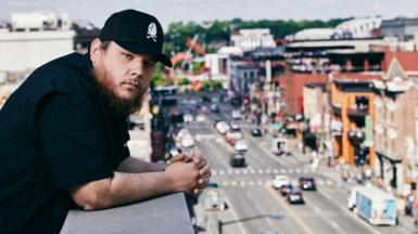In this image released on June 10th, 2021, Luke Combs poses on set for the 2021 CMT Music Awards in Nashville, Tennessee. He is dressed in black with a baseball cap and is overlooking a busy street.