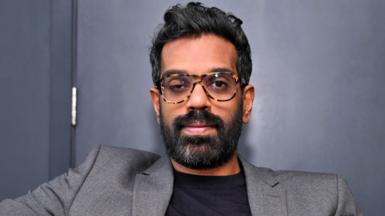 Romesh Ranganathan pictured in front of a navy blue wall, looking at the camera, in March 2024