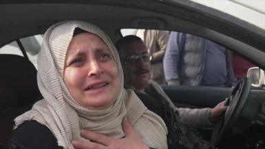 A middle-aged Syrian women is seen sat in the passenger seat of a car. She is wearing a cream-coloured hijab and leaning out the window to talk to a reporter.  She has her left hand on her chest and tears in her eyes. 