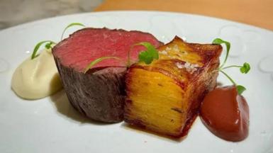 A piece of red meat on a white plate. It is sat beside a potatoe dish. There are dollops of sauce beside the pieces of food on the plate.