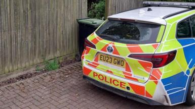 A police car, parked by a fence, with a wheelie bin by it and parked on a brick path. 