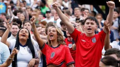 England fans celebrate in a pub garden. A crowd of men and women, in the foreground are two women and two men with their arms up wearing football t-shirts.