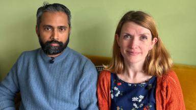 A man, Sree Kadalayil, is on the left and a woman, Jude Pollock, is on the right. They are sitting on an orange sofa that is placed up against a light green wall. Sree has short greying black hair and a black beard with specks of grey. He is wearing a blue jumper. Jude has shoulder-length reddish-blonde hair and is wearing a blue top with a flower pattern and an orange cardigan. 