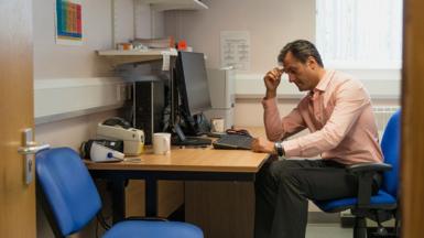 A GP sits at his desk next to an empty patients chair in his surgery, seen through an open door. He is wearing a shirt and dark trousers and is looking thoughtful.