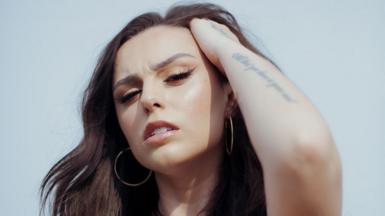 A woman pictured against a cloudless blue sky. She's looking slightly downwards at the camera, giving attitude. Her left hand is placed on her head, pulling back her long black to reveal large, thin gold hooped earrings.  