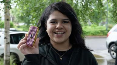 Grace, wearing a black top and jumper, smiles as she holds up her brick-phone, which she has decorated in pink and yellow plastic gems