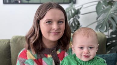 A woman sits with her baby son. She is smiling at the camera. Both are wearing knitted jumpers. A Swiss cheese plant and a green sofa are behind them. 
