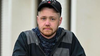 A man in his 30s looks straight at the camera. He is wearing an outdoor jacket, hooded top and is wearing a baseball cap branded 'Zavetti'. He has a neat ginger beard and goatee. His hair is shaved short. 