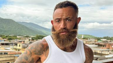 Ashley Cain in Rio de Janeiro in Brazil. He is leaning on a balcony and is wearing brown cargo shorts and a white vest.