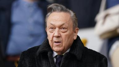 Former West Ham United chairman David Sullivan in the stands before the team's Premier League match at Molineux, Wolverhampton. He is wearing a black coat and tie with a white shirt.