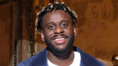 Myles Smith, a man, wearing a white tshirt with a blue denim jacket over the top. He is smiling, with the walls behind him a light brown colour illuminated in some light.