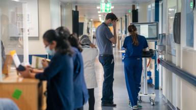 A view of a hospital corridor with various members of staff standing around - two look at a computer monitor
