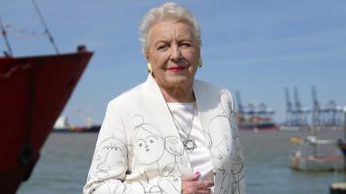 Dame Stephanie Shirley poses infront of the sea in a white suit covered in drawings of children and a Star of David necklace at the unveiling of a memorial to Kindertransport on the quayside at Harwich in 2022