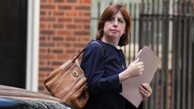 Lucy Powell is holding a brown folder and has a large brown-coloured bag over her right shoulder. She is wearing a dark blue jacket. A brick wall and a metal fence can be seen in the background.