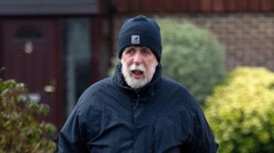 A grey bearded Gareth Jenkins wearing a dark blue beanie hat and dark blue coat, walking in front of a house