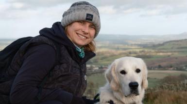 Natalie in grey beanie in dark hoodie and body warmer with golden retriever dog stood in front of hillside landscape