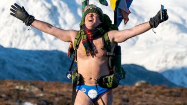 Speedo Mick Speedo Mick wearing his blue Speedos, a woolly hat, scarf, backback, gloves and nothing else, his face tilted up[wards to the sky with his eyes closed and a look of relief, against a background of snowy mountains in Glencoe, Scotland, in January 2023