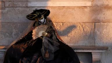 A masked reveller sits on a stone bench beside a brick wall 