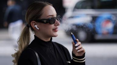 A young woman wearing a tracksuit top, Prada sunglasses and Airpods uses a blue-coloured disposable vape on a London street