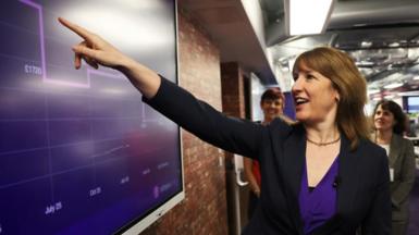 Rachel Reeves smiles while pointing to a large purple chart on a screen, which shows Octopus energy prices and months. The chart appears to show prices falling.