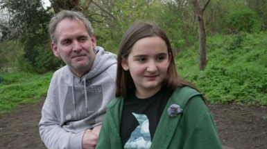 A girl in a green jacket and a penguin top sits on a bench with her dad who is dressed in a grey jumper.