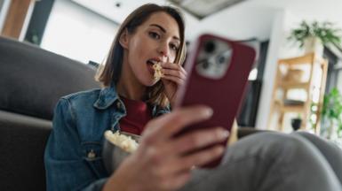 A woman eating popcorn from a bowl while scrolling on her red smartphone