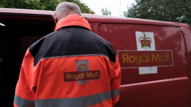 An employee of postal service Royal Mail sorts parcels and letters in the back of his red delivery van baring the postage company's signage. He wears an orange high-vis jacket baring the same logo.