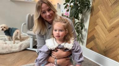 A young girl in a lilac top with white collar being cuddled by her mum, who has long blonde hair and is wearing a grey top