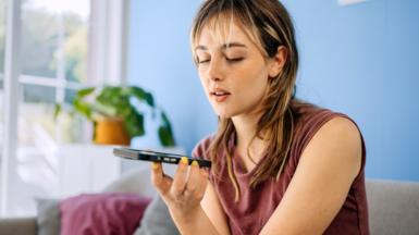 A young woman casually seated on a sofa, holding her smartphone and sending a voice message.
