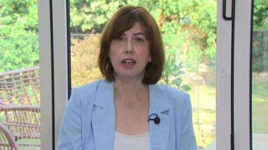 Lucy Powell wearing a blue jacket and a white top. She is sitting in front of windows that look out on to a garden.