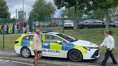 Outside shot of the school where it took place. A parked police car is seen , in front of the school gates. Two people walk past.