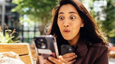 woman with long dark hair looks at her phone, with an expression of joy on her face.