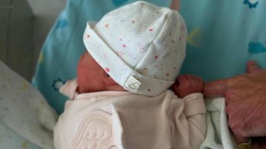 A baby wearing a pink sleepsuit and a white hat with pink decoration. The finger of an older adult's hand is in the picture with the baby clasping it