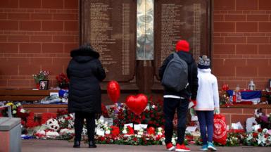People stand in front of the Hillsborough Memorial outside Anfield Stadium in Liverpool. There are flowers and heart balloons in front of a plaque with the names of the 96 victims of the disaster.