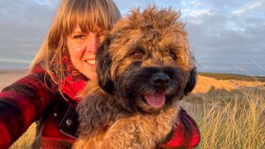 A woman in a red jacket cuddles a small, dog. The dog is looking into the distance and has its tongue out and looks happy. There is a beach in the background and it is a sunny day. 