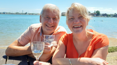 Chris and Ruth Stone-Houghton sit on a picnic bench, smiling into the camera. They are holding alcoholic drinks, and behind them is a body of water